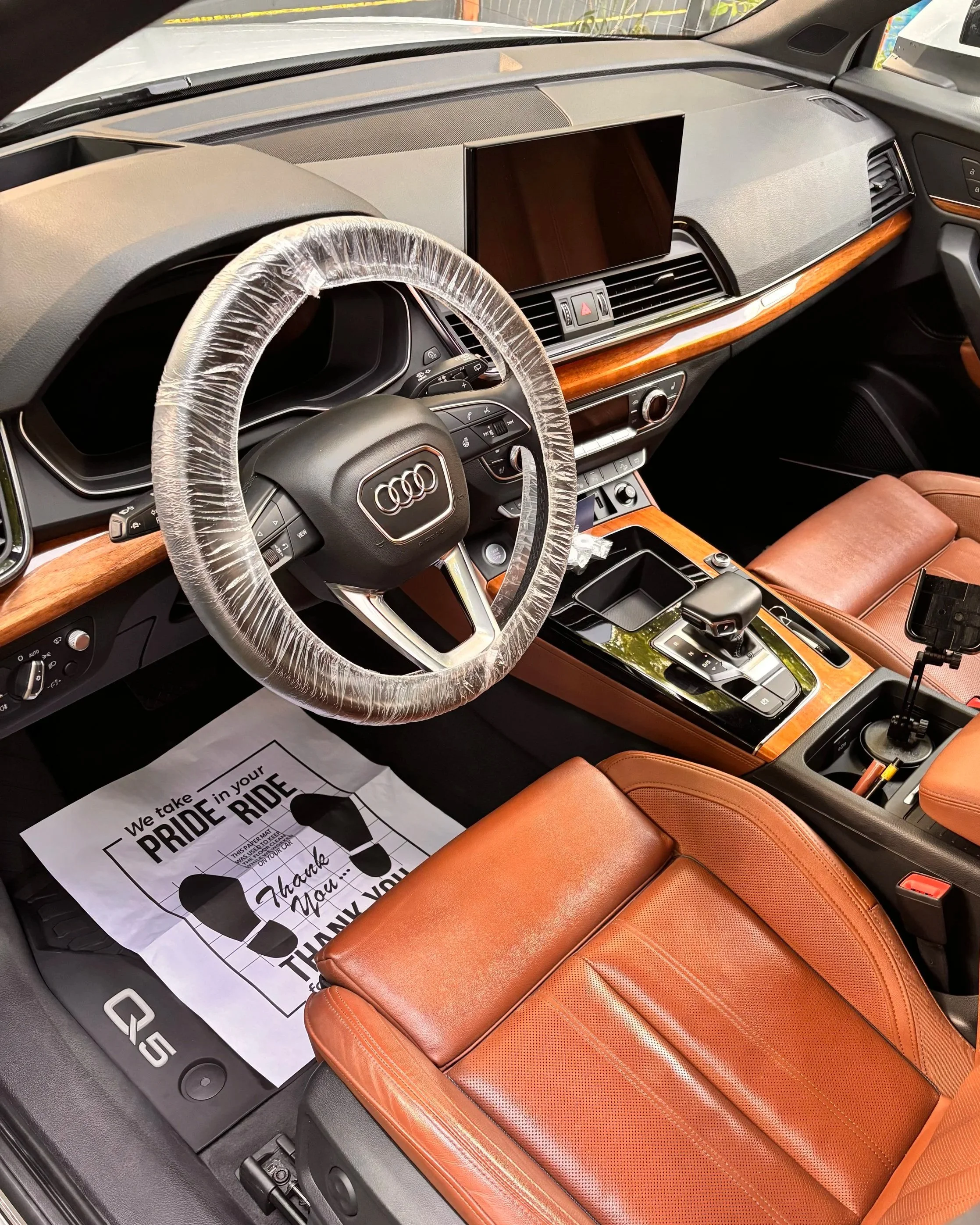 Interior of an Audi Q5 SUV with a leather seat, wooden dashboard accents, a large touchscreen display, and a steering wheel wrapped in plastic.