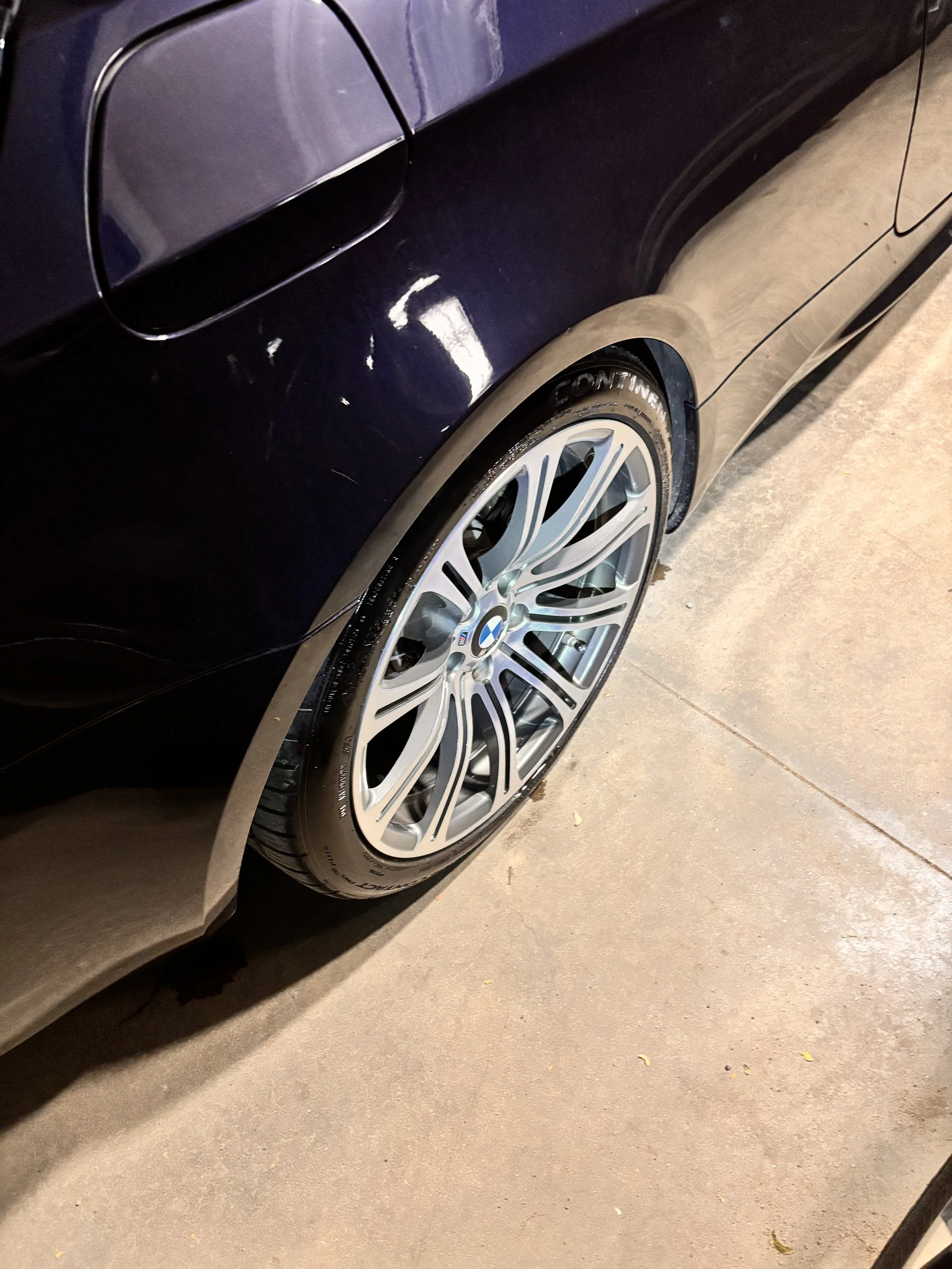 Close-up of a dark blue BMW car's rear left side, showing the wheel with a low-profile tire and the body with minor scratches.
