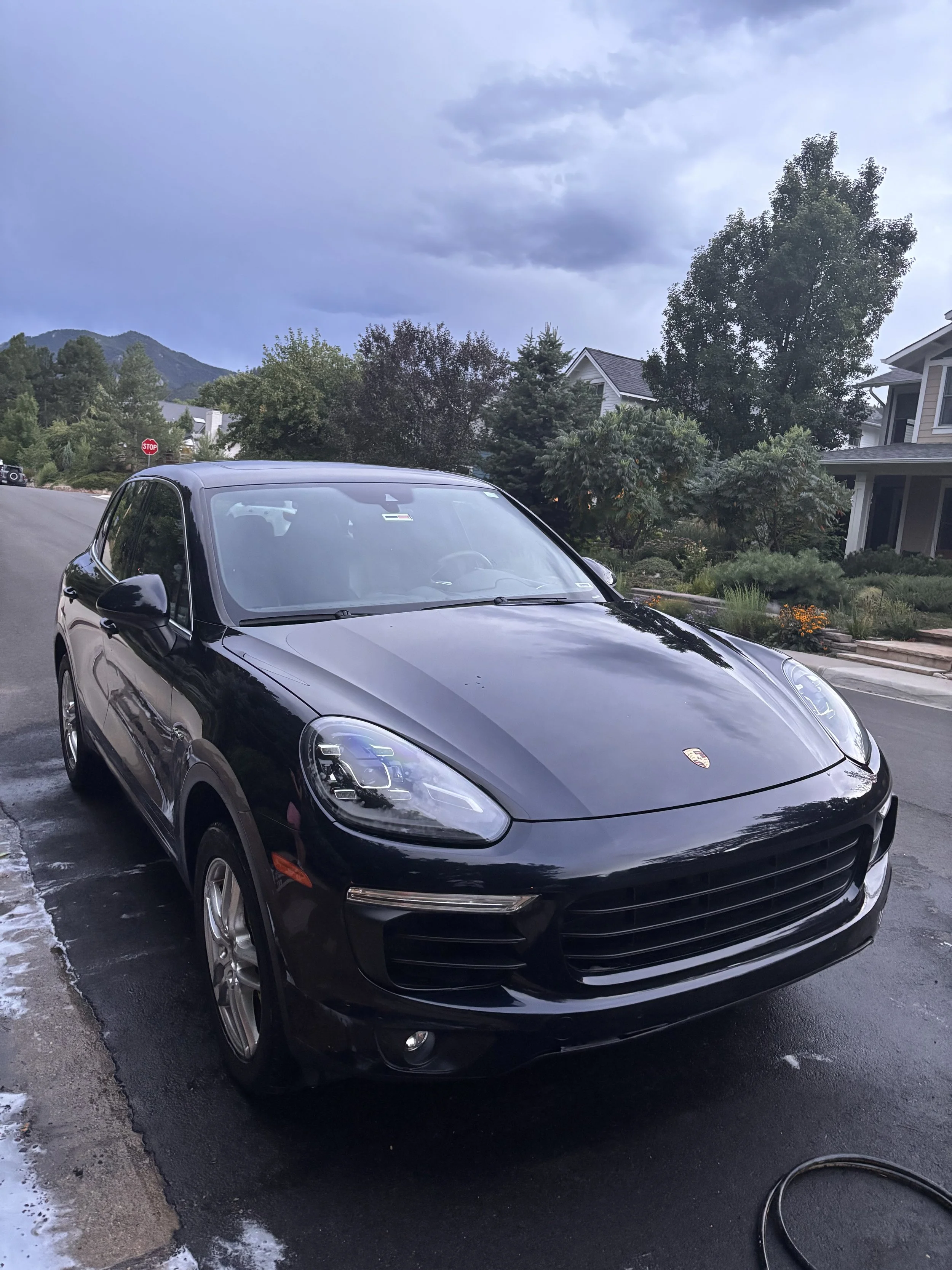 Black Porsche SUV parked on a driveway in a suburban neighborhood with trees and houses under a cloudy sky.