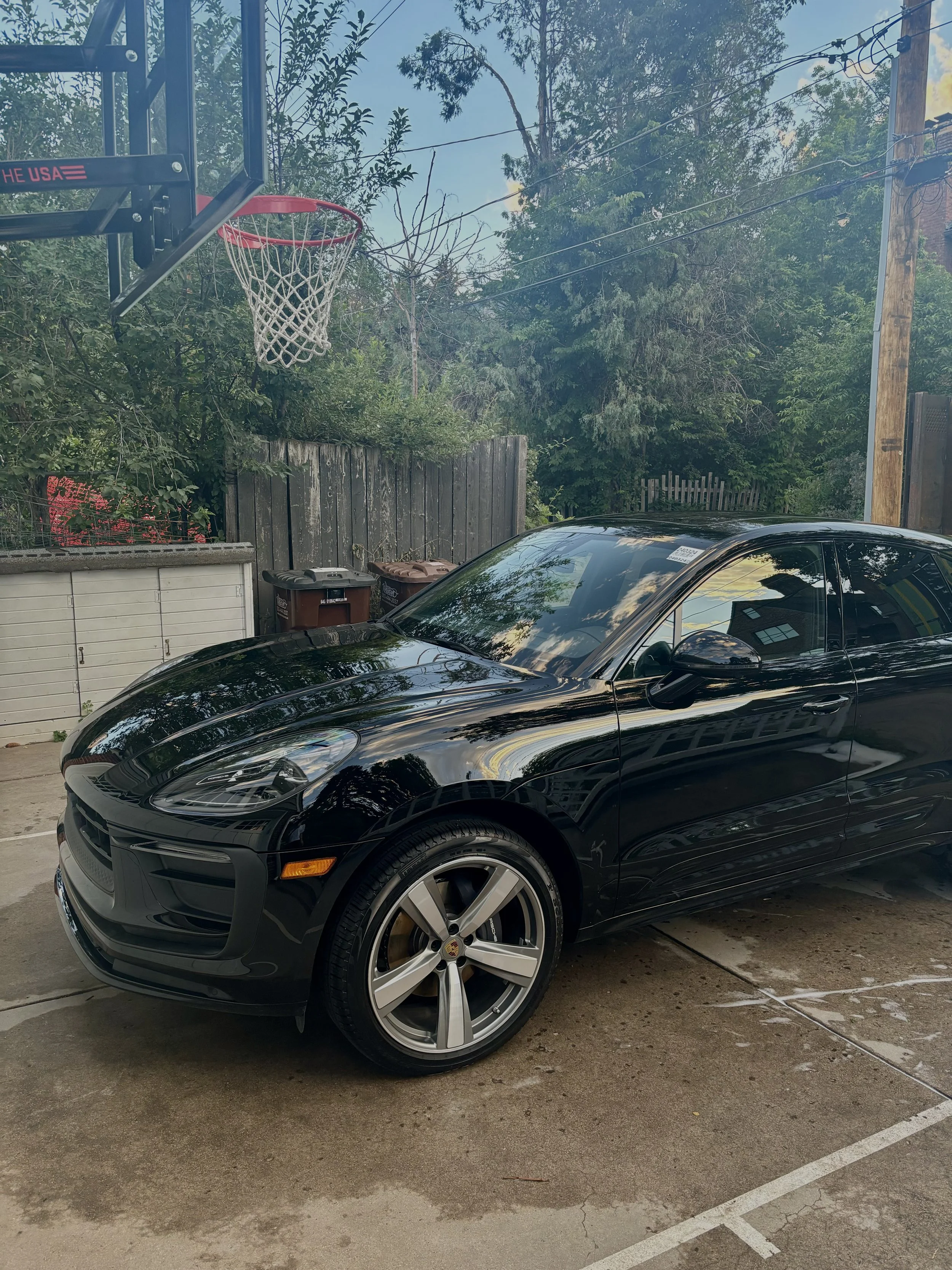 A black sports car parked in a driveway with a basketball hoop overhead. There is a wooden fence and trees in the background.