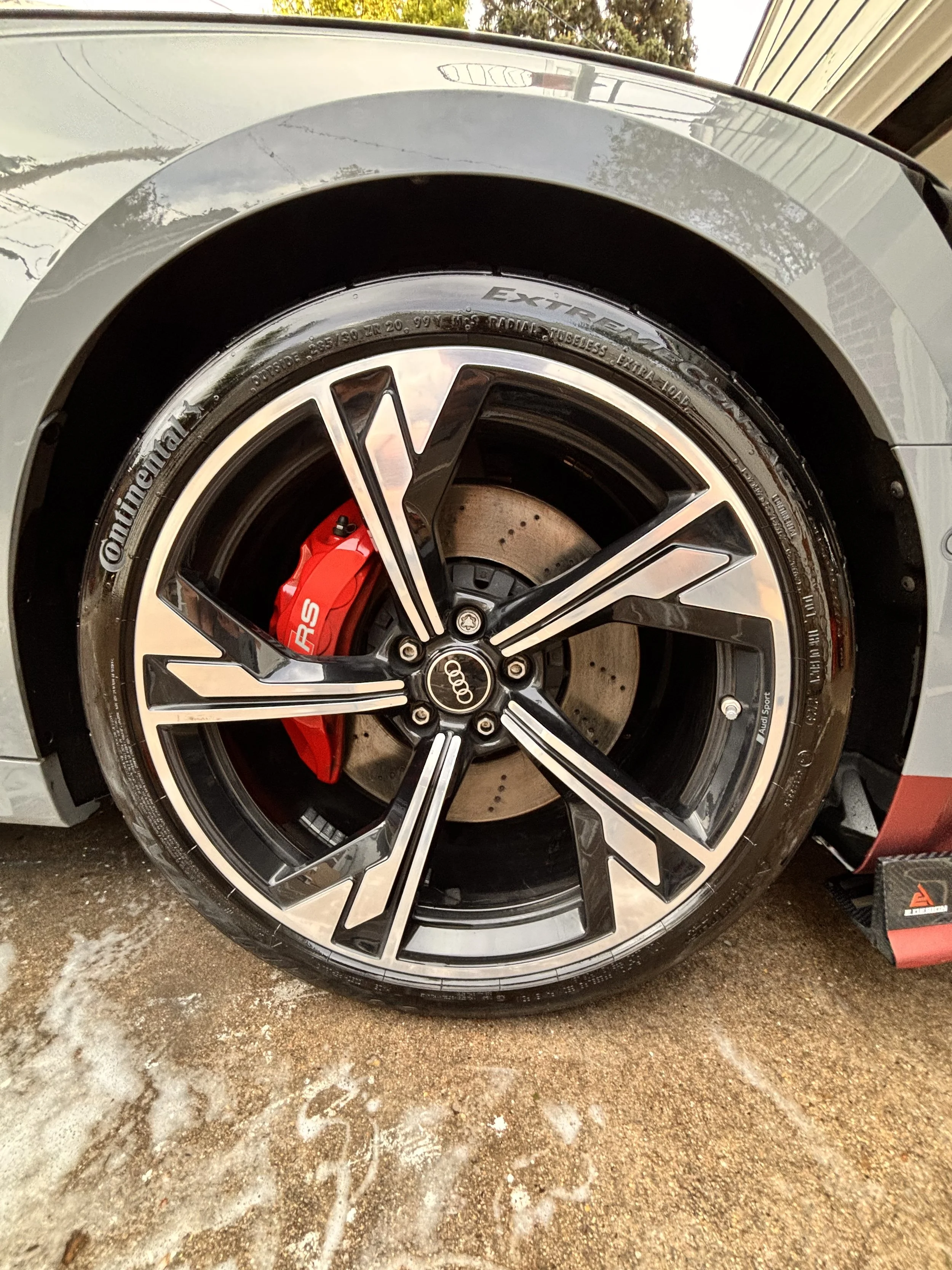 Close-up of a car wheel with a black and silver rim, red brake caliper, and drilled brake disc, attached to a silver car.