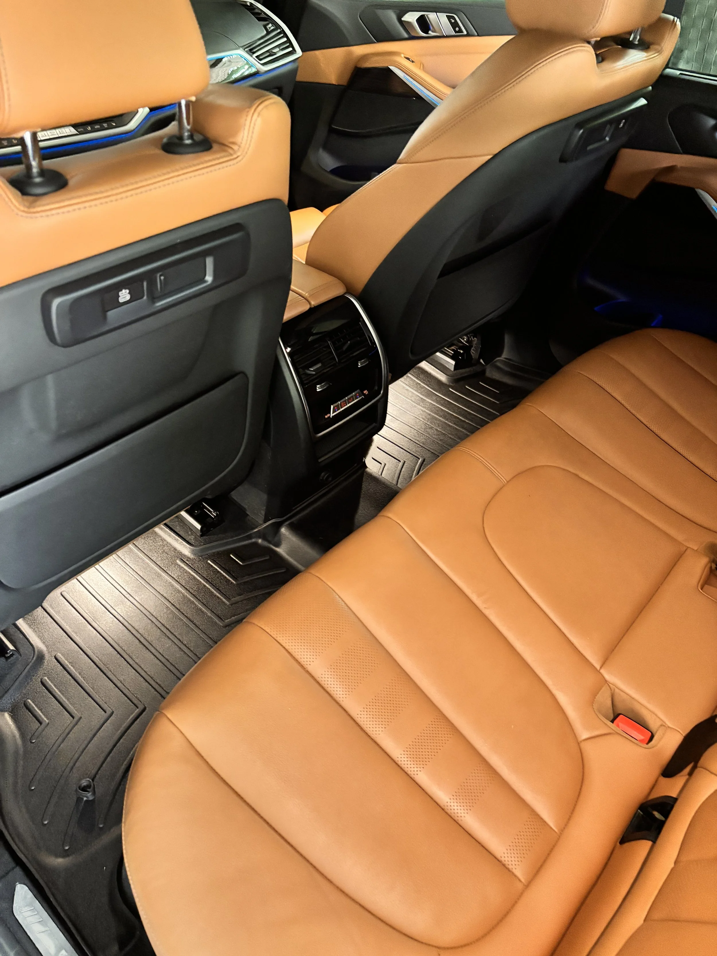 The interior of a car showing tan leather seats, black floor mats, and the back of the front seats with built-in controls. Air vents and climate control are also visible between the seats.