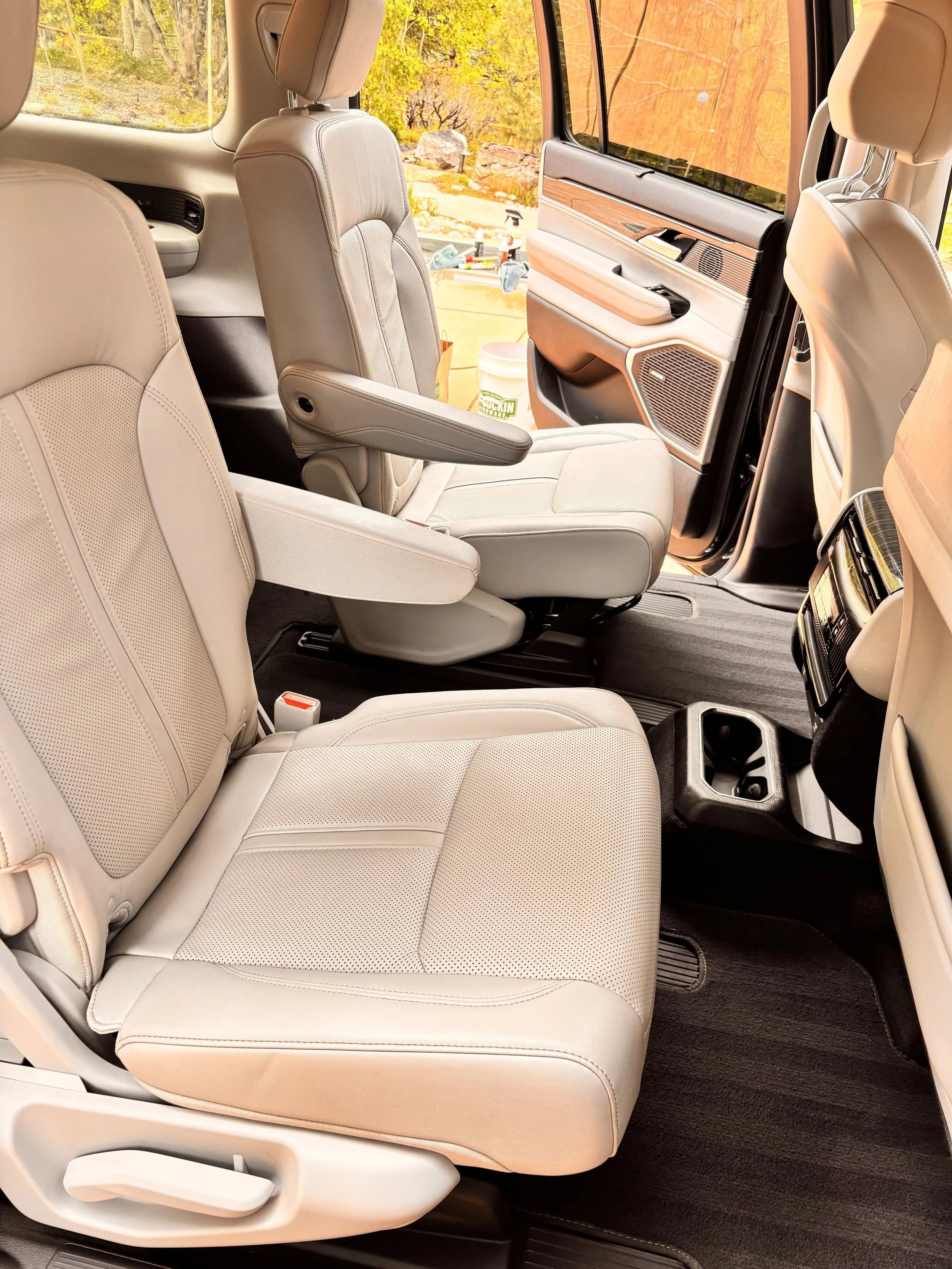 Interior of a vehicle showing beige leather seats, with the front passenger seat and rear seats visible. The vehicle door is open, revealing a sunny outdoor scene with trees and items outside.