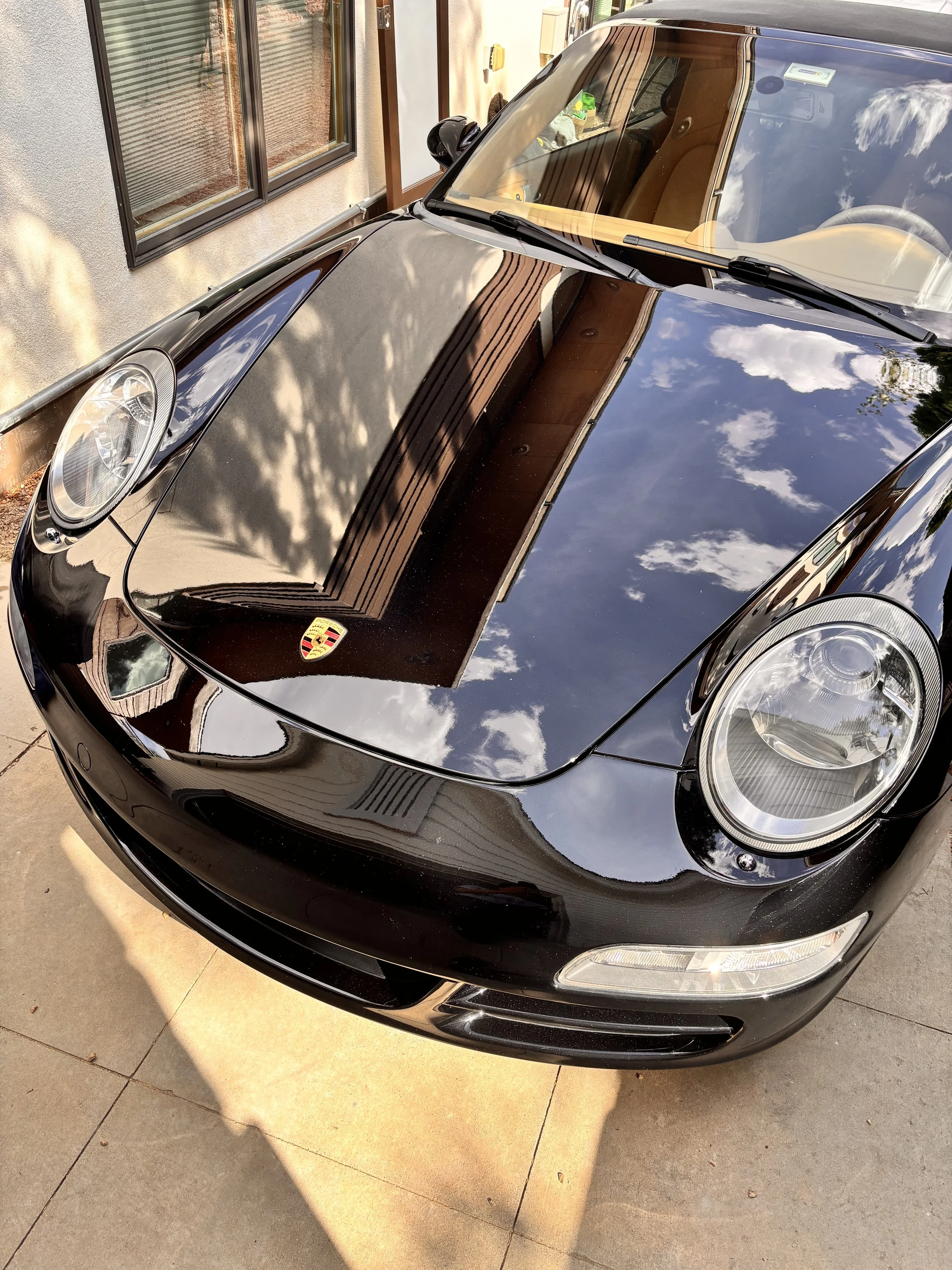 Black Porsche sports car parked on a driveway, reflecting the sky with clouds and nearby house. The Porsche badge is visible on the hood.