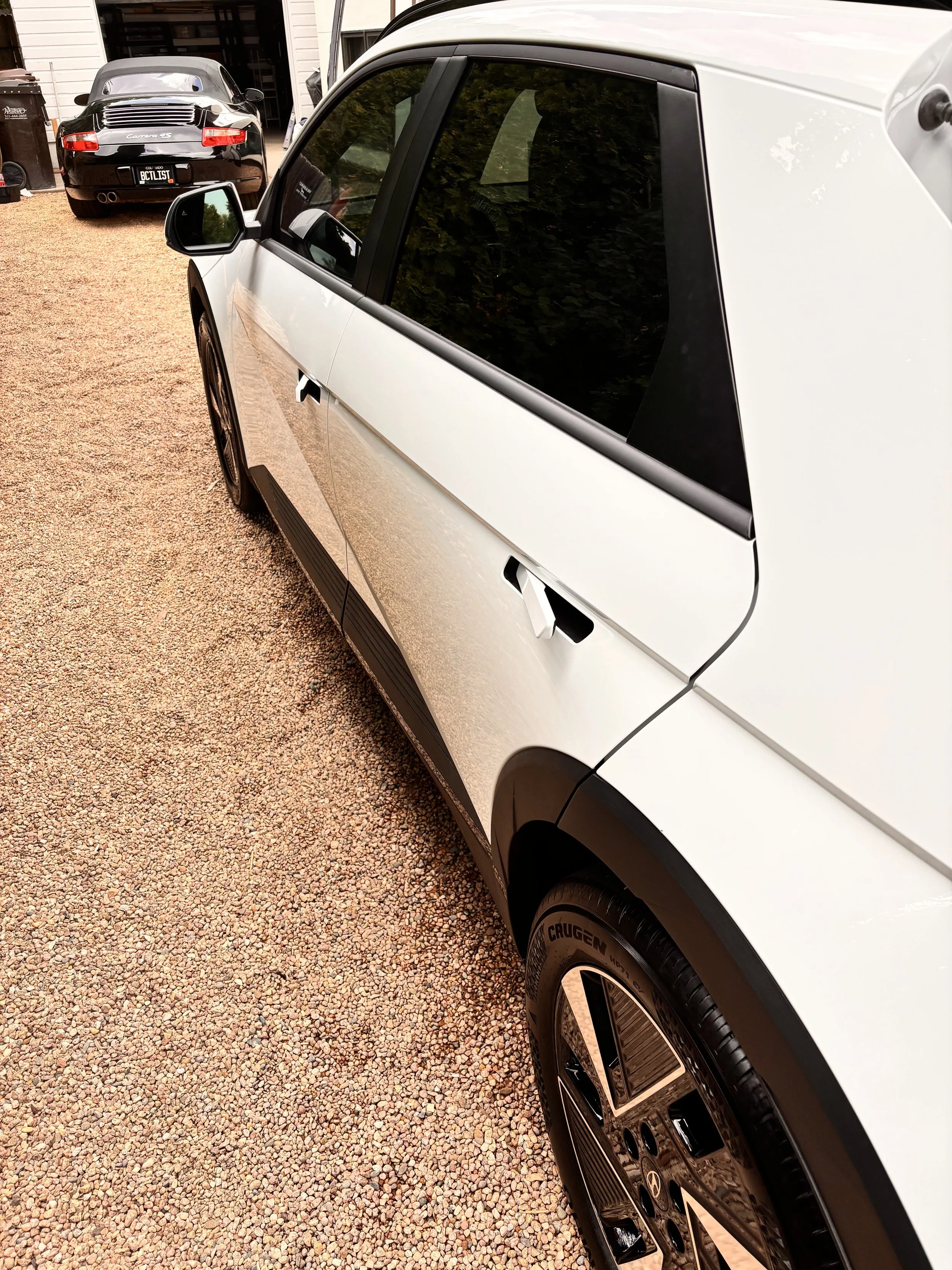 A white and black modern SUV parked on a gravel driveway, with another black sports car and a garage in the background.