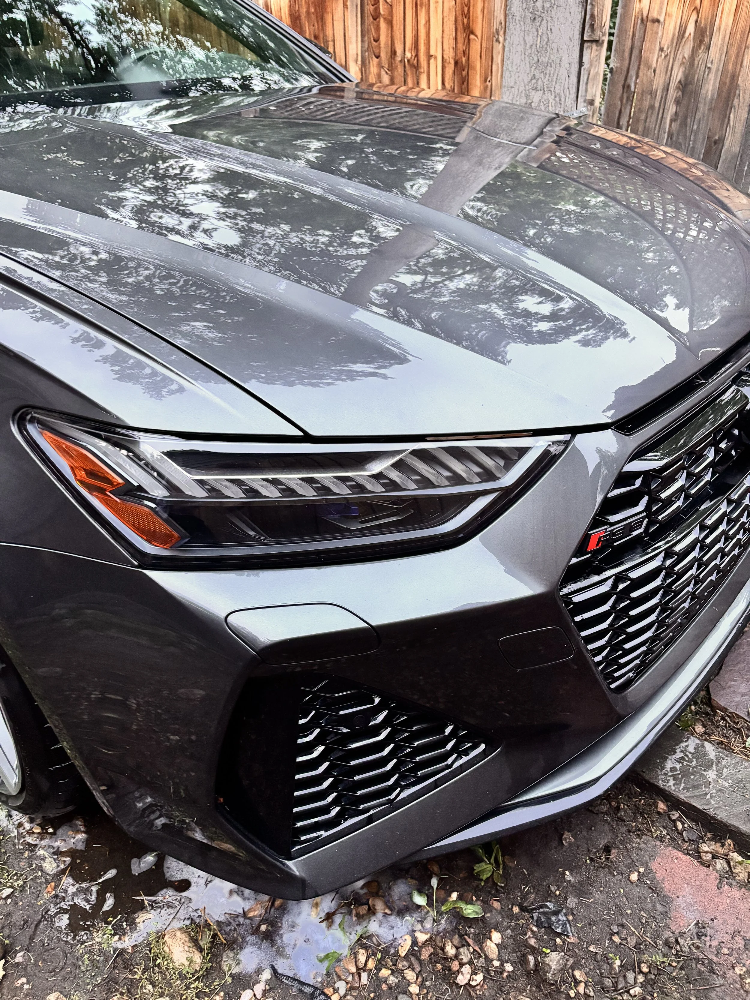 Close-up view of the front of a gray Audi car parked outdoors, showing the headlights, grille, and part of the hood reflecting trees and the sky.