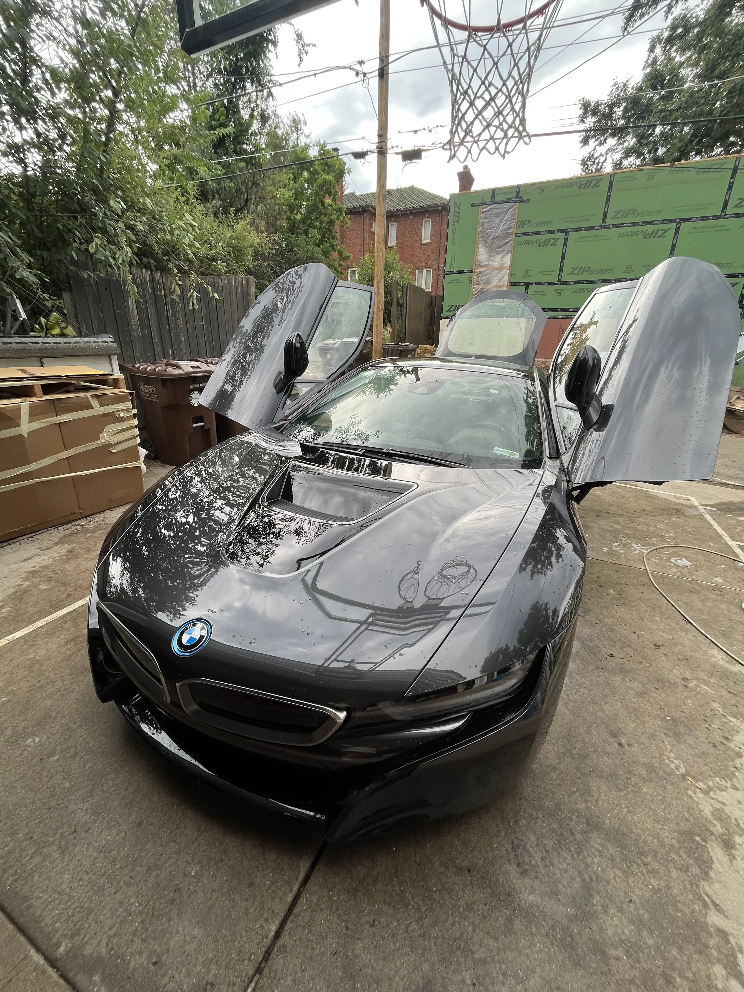 A black BMW sports car with butterfly doors open, parked in a driveway with a basketball hoop overhead. There are trees, a fence, and a building with green insulation boards in the background.