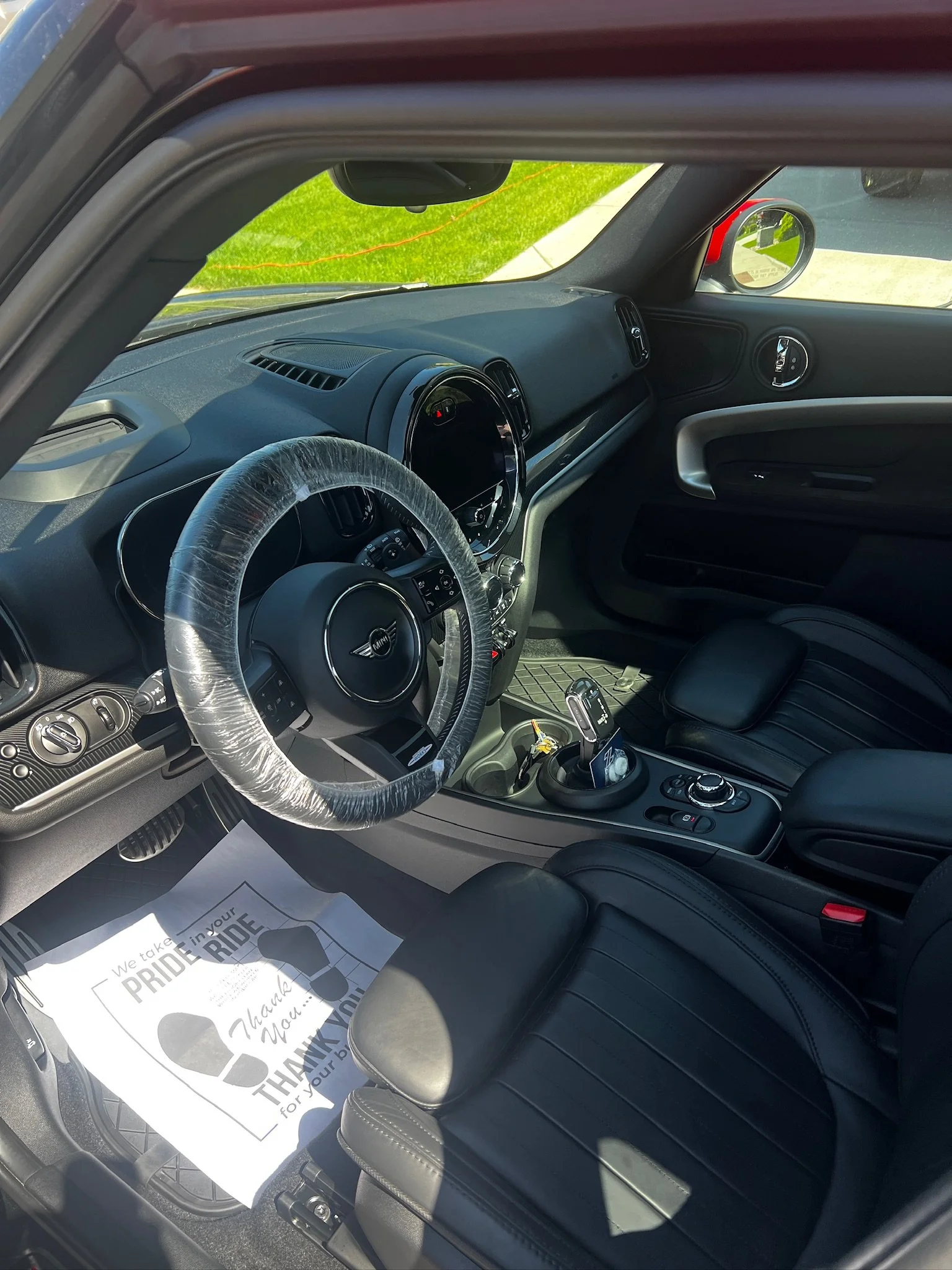 Inside view of a car with a black interior, plastic-wrapped steering wheel, gear shifter, and a paper on the floor with footprints and thank you message.