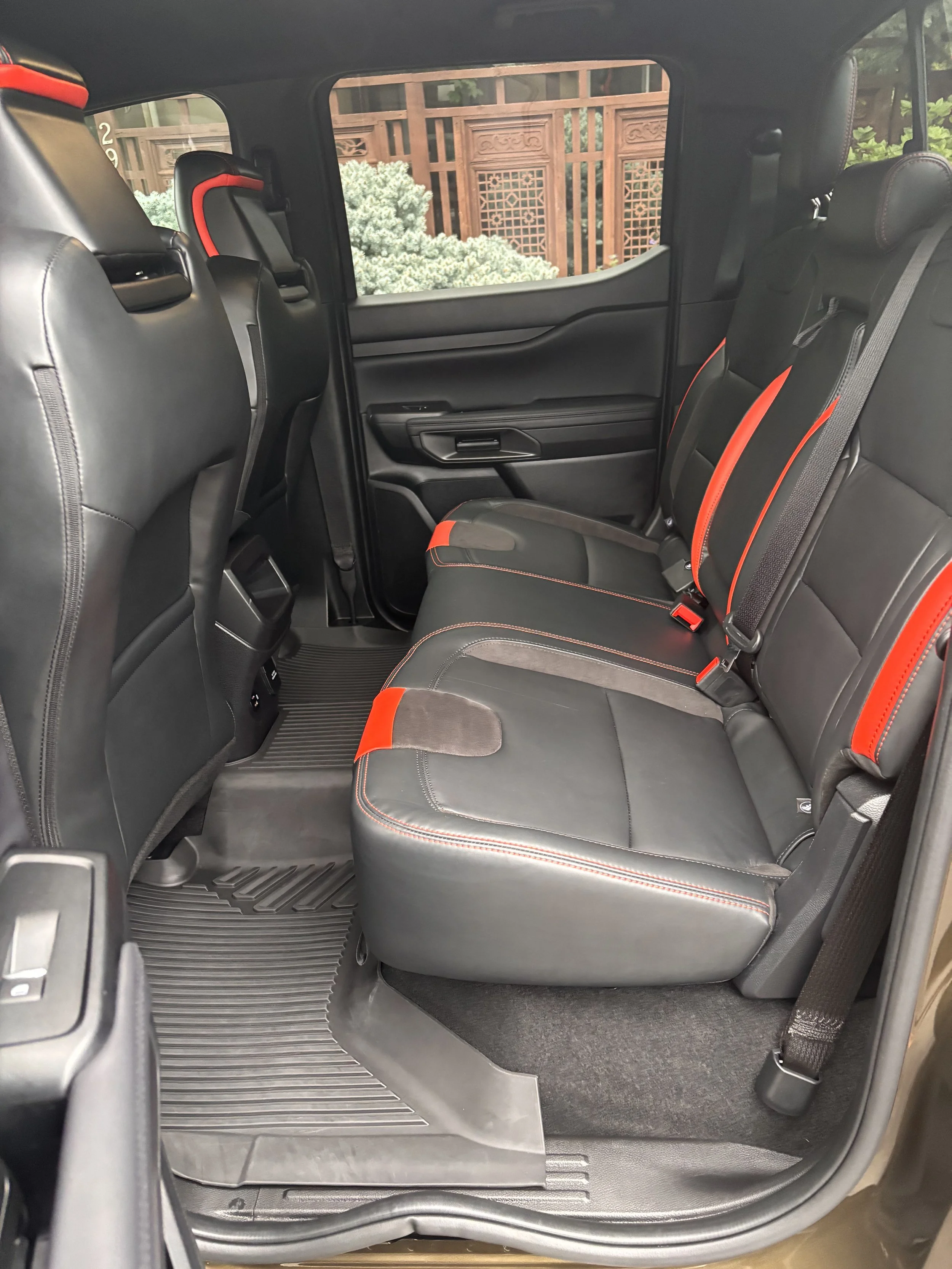 The interior of a vehicle showing three black leather seats with red accents, seatbelts, and rubber floor mats, with a window revealing a wooden fence and some greenery outside.