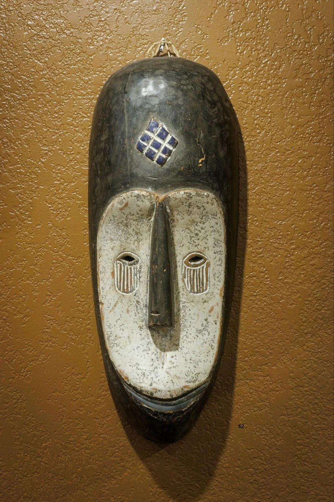 A traditional African tribal mask with an elongated face, black and white colors, and a geometric design on the forehead, hanging on a brown textured wall.
