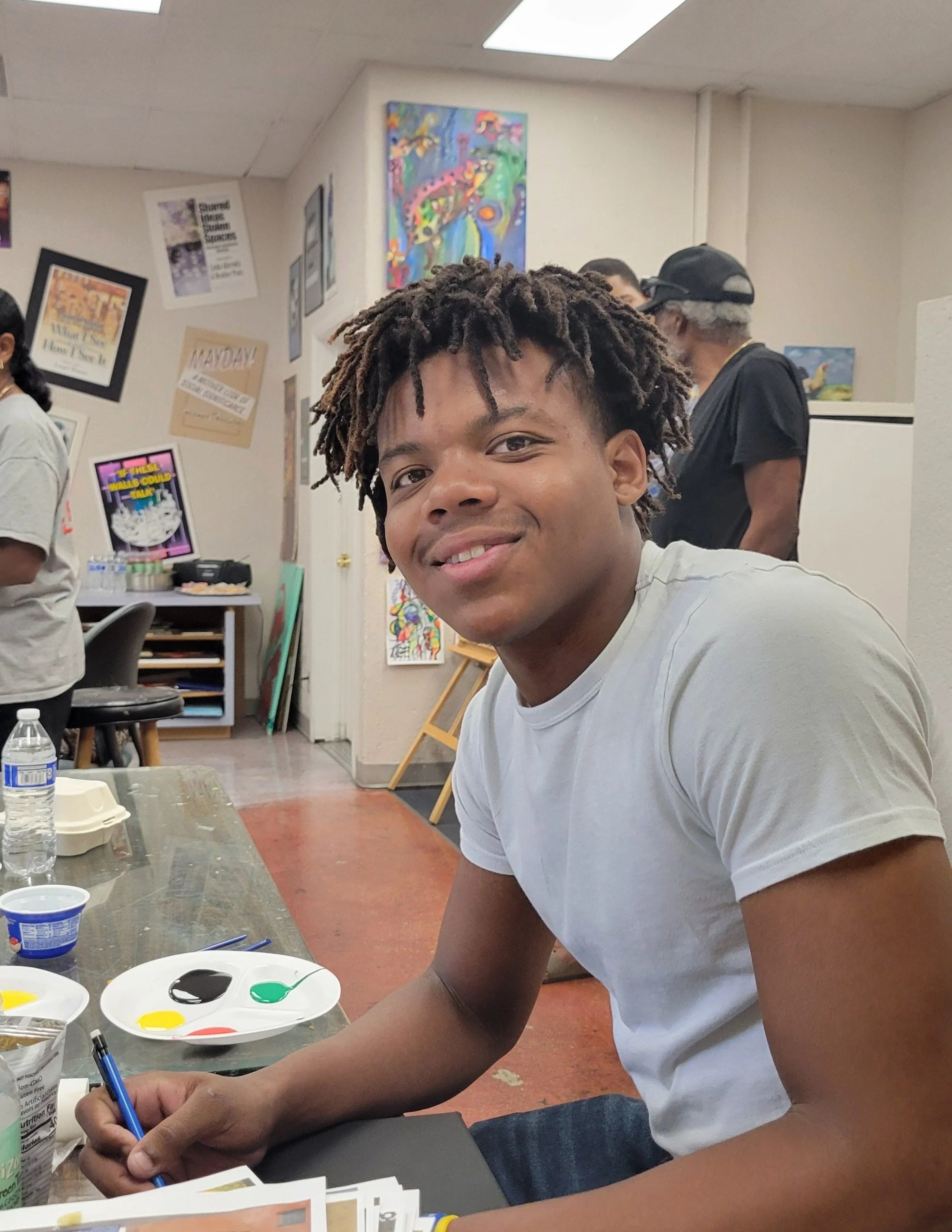 Young man with dreadlocks and light-colored t-shirt smiling at camera while sitting at a table with paint supplies, in a room decorated with colorful artwork and posters.