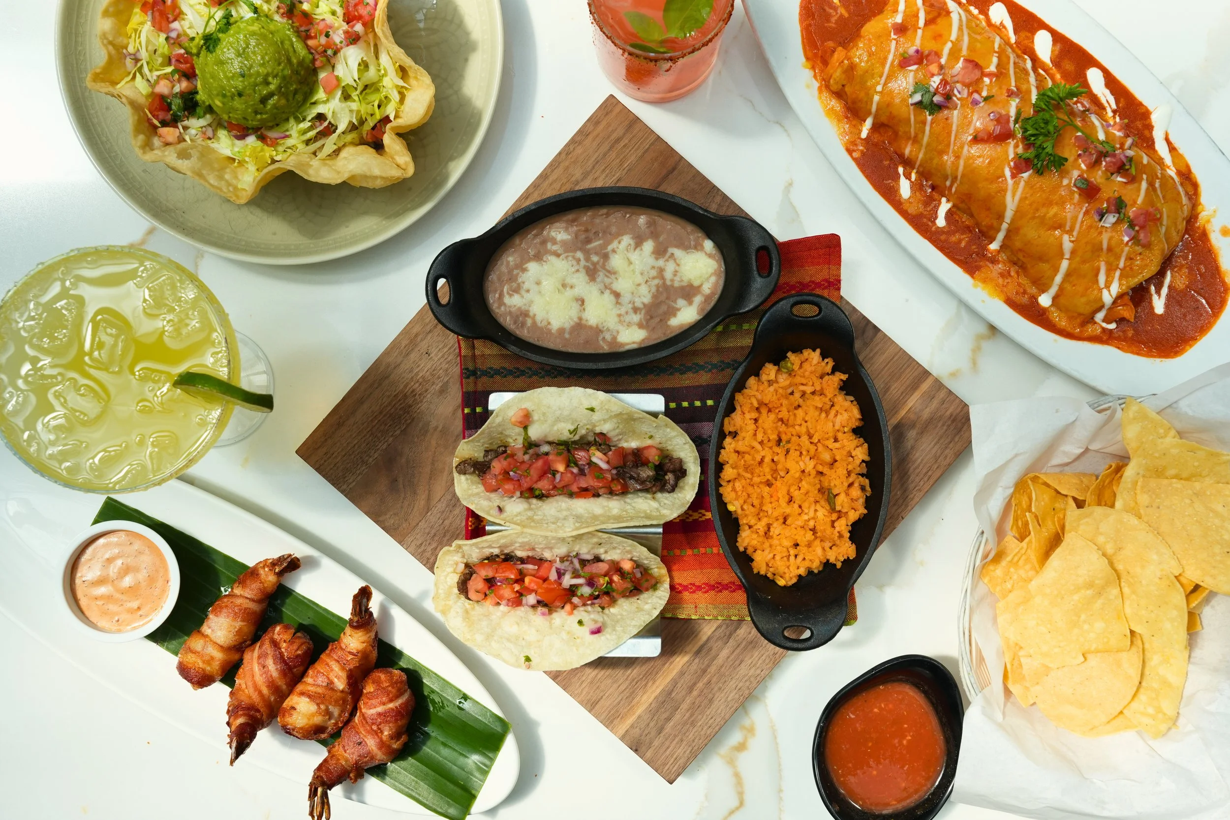 Assorted Mexican dishes including tacos with diced tomatoes and onions, rice, beans, a burrito smothered in sauce, nachos, bacon-wrapped shrimp, a side of chips with salsa, and beverages including lemonade and a pink drink.