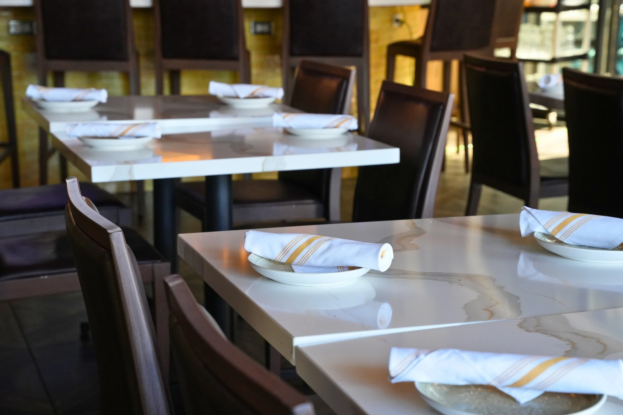 Empty restaurant dining tables set with plates and napkins, with dark wood chairs.