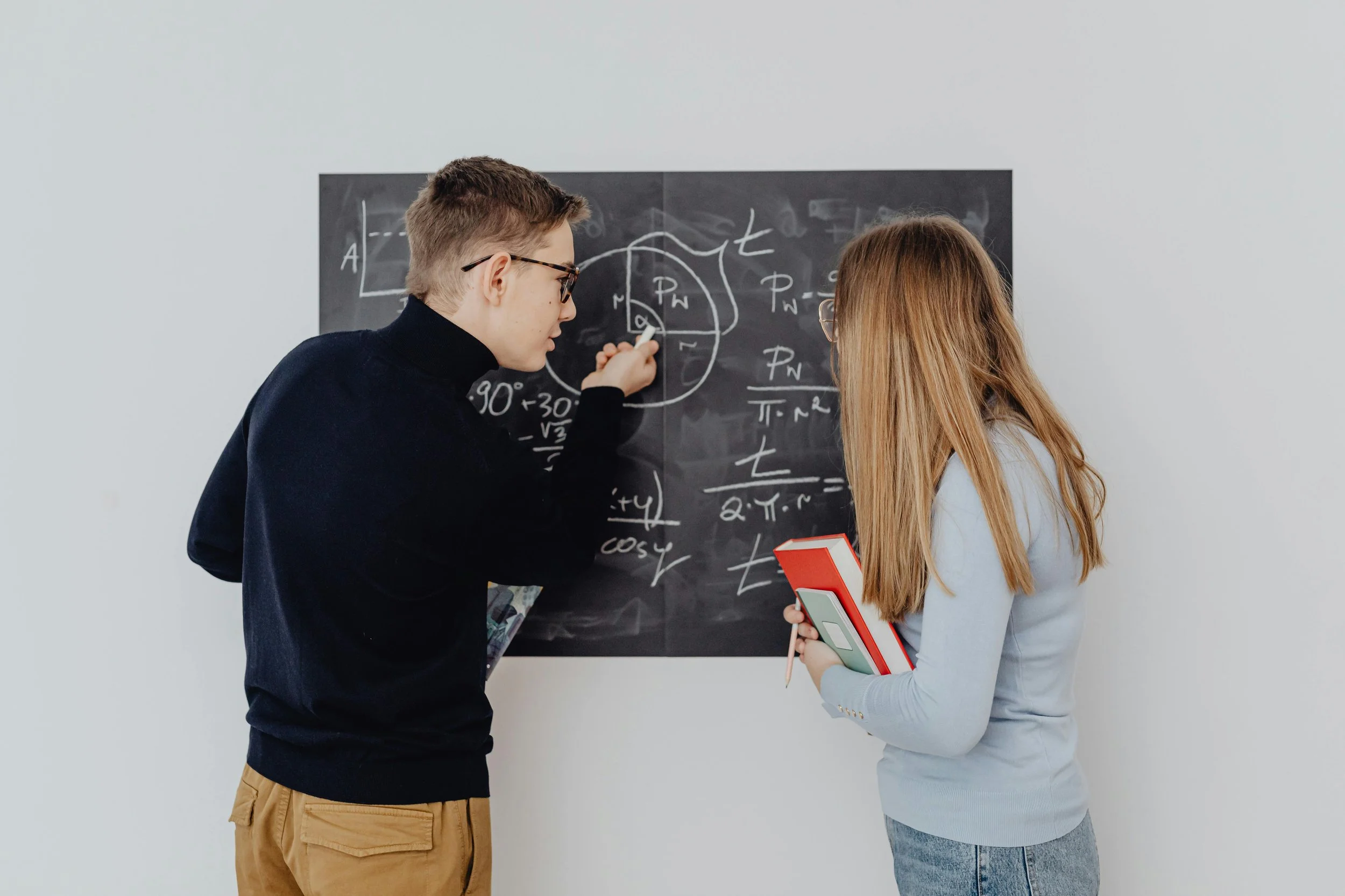 A young man and woman discussing physics concepts in front of a blackboard filled with equations and diagrams, with the young woman holding a folder and a notebook.