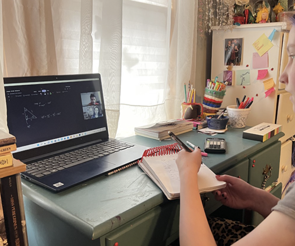 A student sitting at a cluttered desk engaged in an online class, with a laptop, notebooks, and colorful school supplies, in a room with natural light and colorful decorations on the wall.