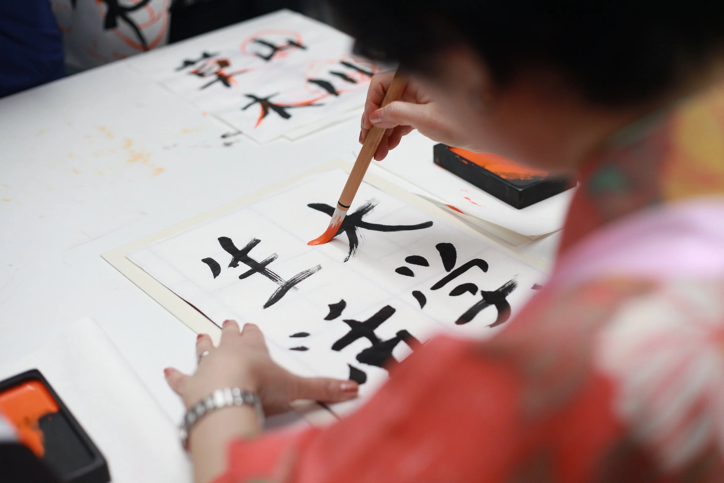 A person practicing Asian calligraphy, writing bold black Chinese characters on a white sheet with a brush dipped in orange ink.