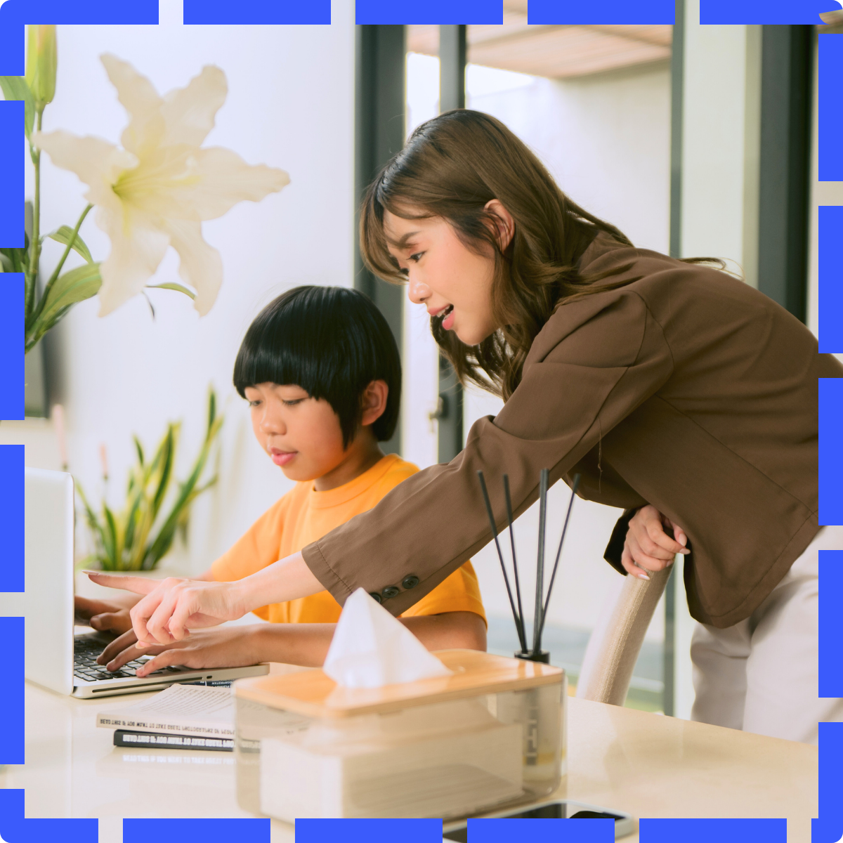 A woman is helping a young boy with a laptop at a table in a bright room with plants and flowers.