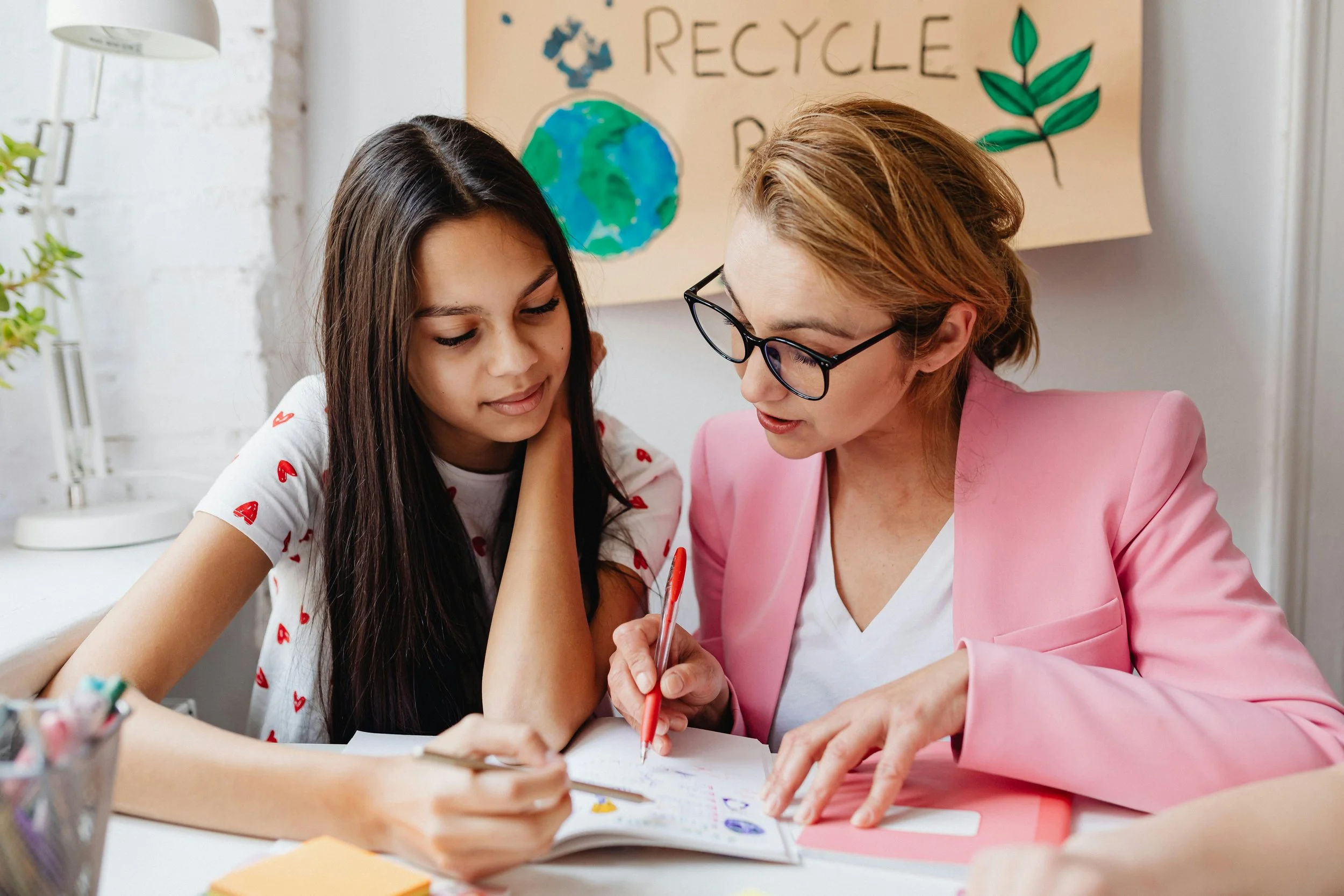 A teacher and a student are sitting at a desk, working on an assignment together. The teacher is pointing to a notebook while the student listens attentively. Behind them, there is a poster with the words 'Recycle' and a drawing of the earth and a plant.