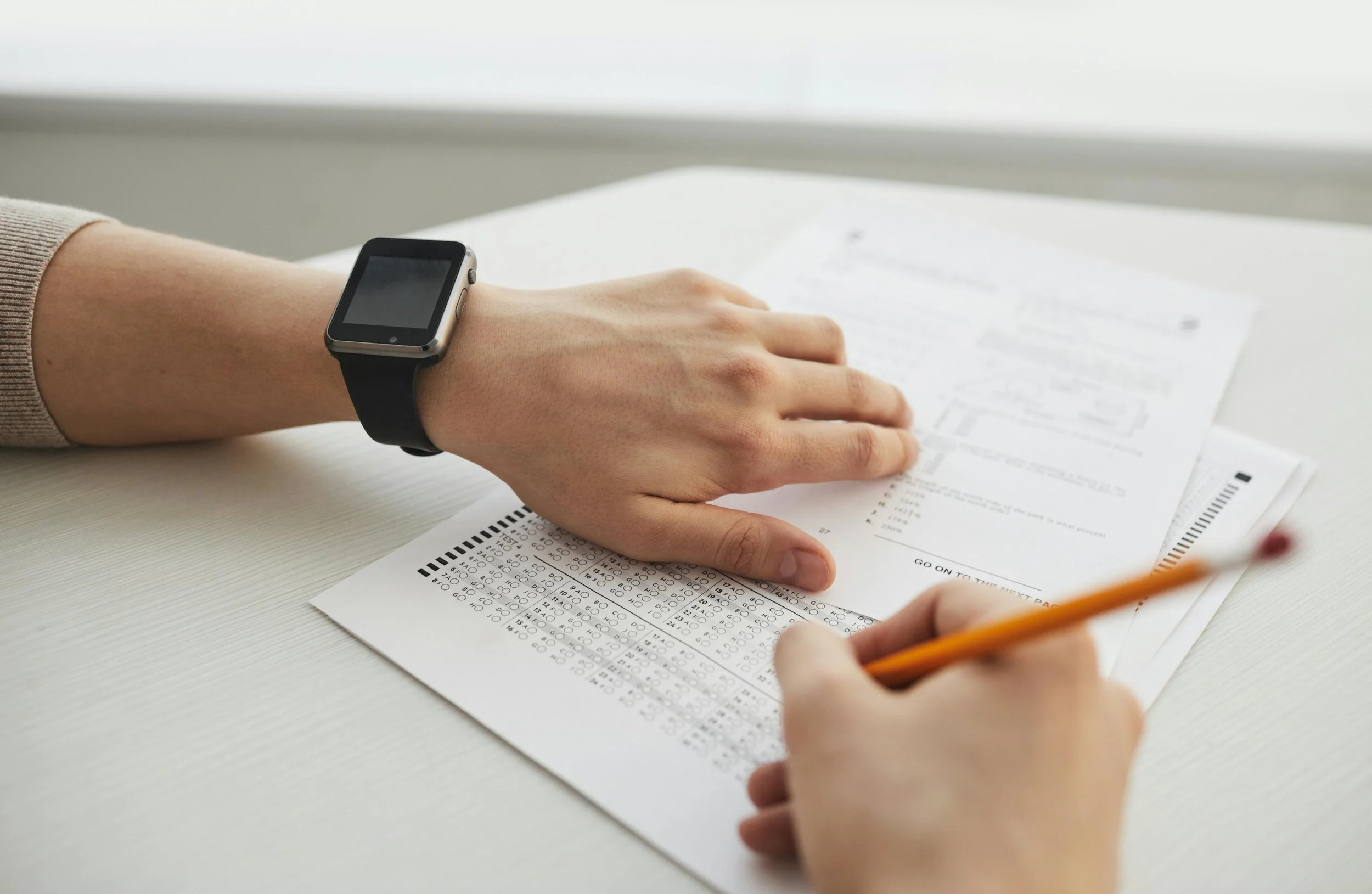 A person is taking a test with a pencil, wearing a smartwatch, and holding a sheet of answer sheet on a white desk.