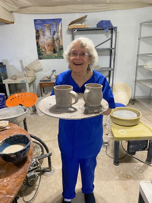Older woman in blue scrubs smiling and holding a pottery wheel with two ceramic mugs on it in a pottery studio.
