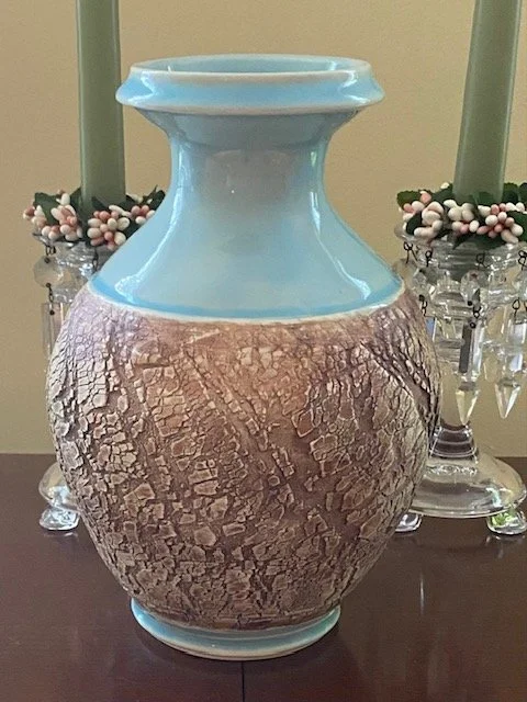 Large decorative vase with a light blue top and a textured, cracked brown bottom, placed on a table with candles and floral arrangements in the background.