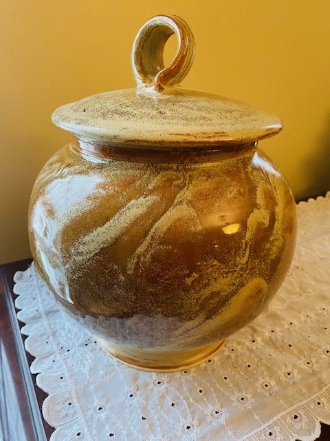 A ceramic jar with a lid and a loop handle, placed on a white lace doily.