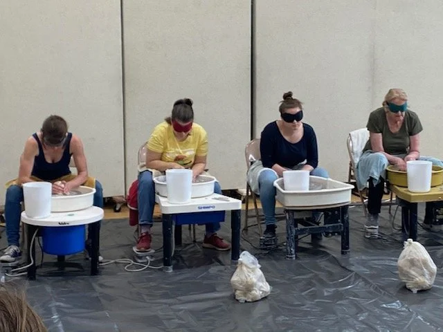 Four women sitting at pottery wheels, each wearing eye protection, working on clay pots in a studio with a black floor and plain light-colored walls.