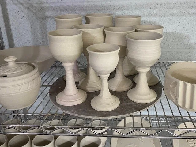 Multiple unfinished white ceramic pieces, including cups and bowls, on a wire rack in a pottery studio.