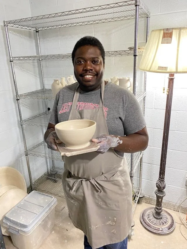 A smiling man in a gray t-shirt and apron holding a ceramic bowl, standing in a pottery studio with metal shelves and a floor lamp nearby.