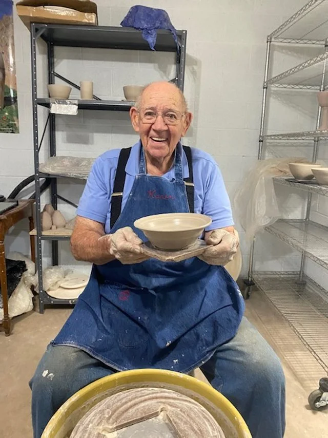 An elderly man wearing a blue apron and gloves, sitting in a pottery studio, holding a finished ceramic bowl and smiling. Shelves with pottery tools and other ceramic items are in the background.