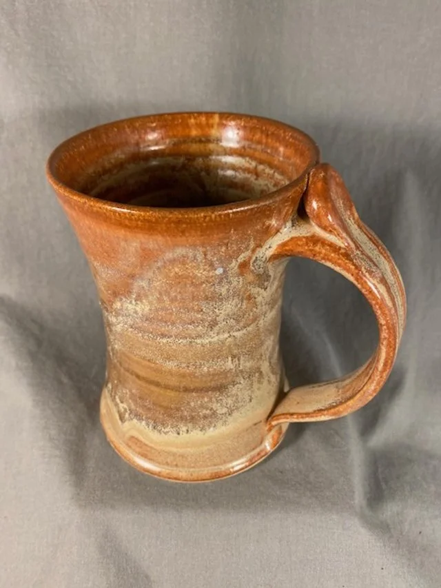 A ceramic mug with a rustic, earthy glaze and a distinctive curved handle, standing on a gray surface.