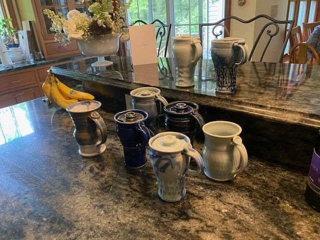 Several small ceramic jugs and mugs on a kitchen countertop with a bunch of bananas and a flower arrangement in the background.