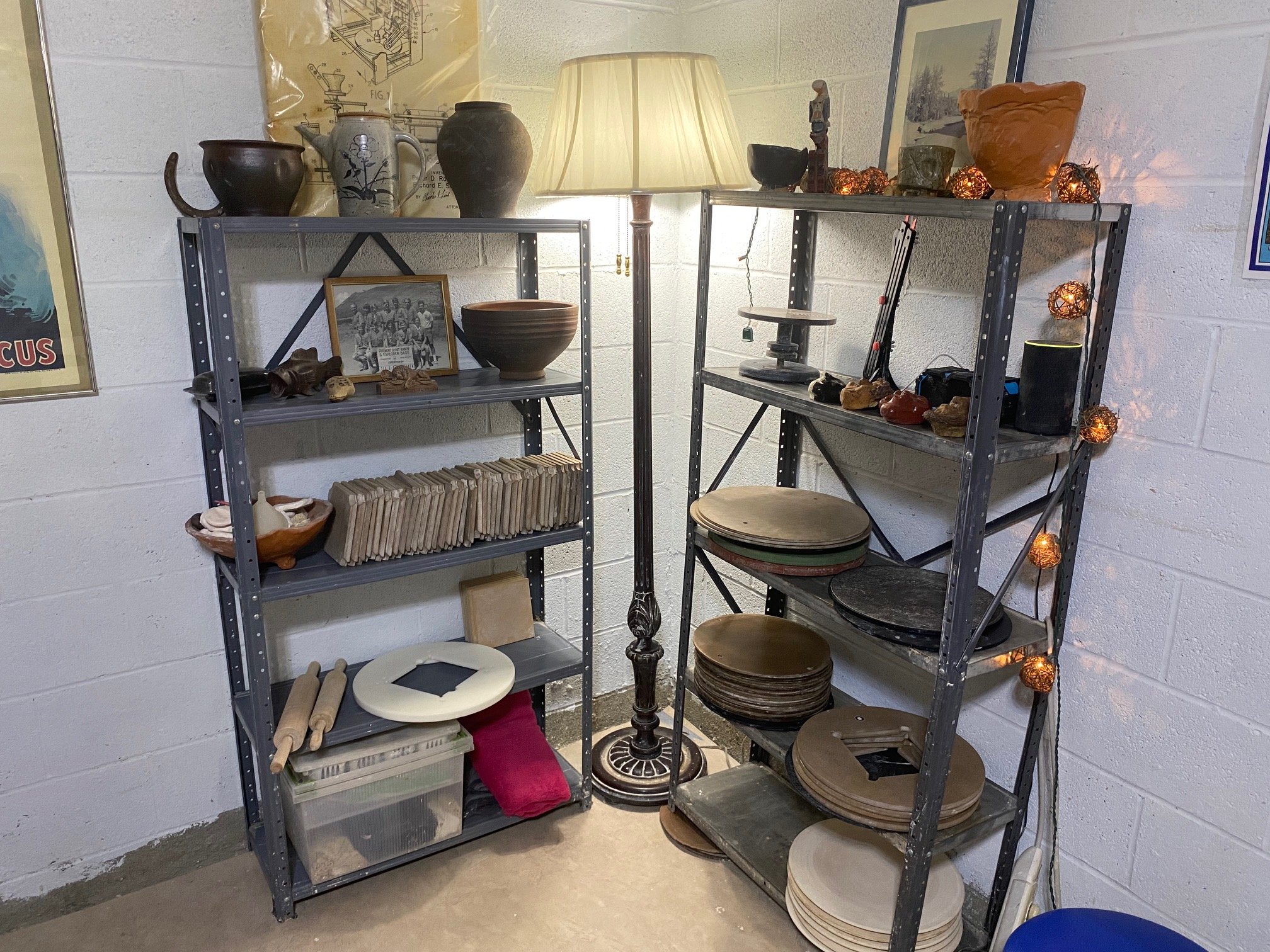 Two metal shelving units filled with pottery, picture frames, and miscellaneous items, illuminated by a floor lamp, against a white brick wall decorated with string lights.