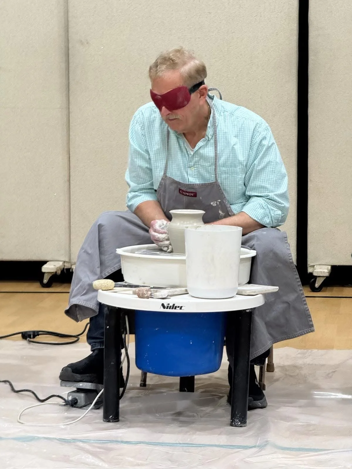 A man wearing a blindfold and apron is sitting at a pottery wheel, shaping clay into a vase.