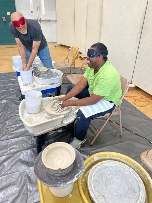Two individuals in a pottery workshop, one sitting and blindfolded, learning to shape clay on a pottery wheel, while the other stands nearby assisting. There are multiple pottery wheels and buckets with clay and water around them.