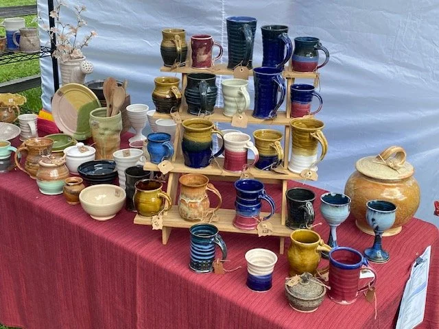 An outdoor display of colorful ceramic mugs, bowls, and pots arranged on a table covered with a red cloth, with some items on wooden shelves.