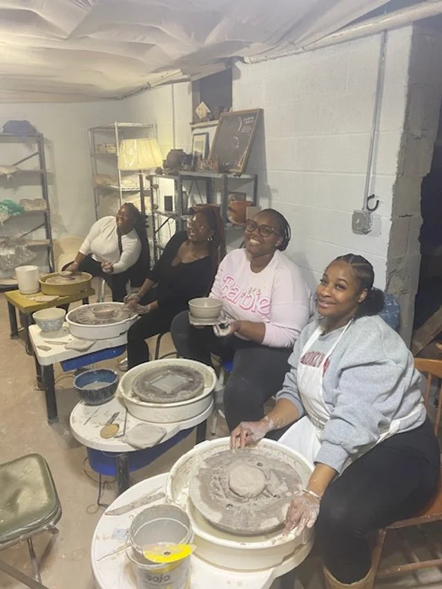 Four women sitting at pottery wheels making clay pottery in a basement workshop, smiling and enjoying their activity.