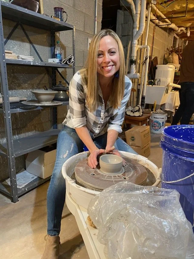 A woman with long blonde hair, wearing a plaid shirt and jeans, smiling while working on a pottery wheel in a ceramic studio.