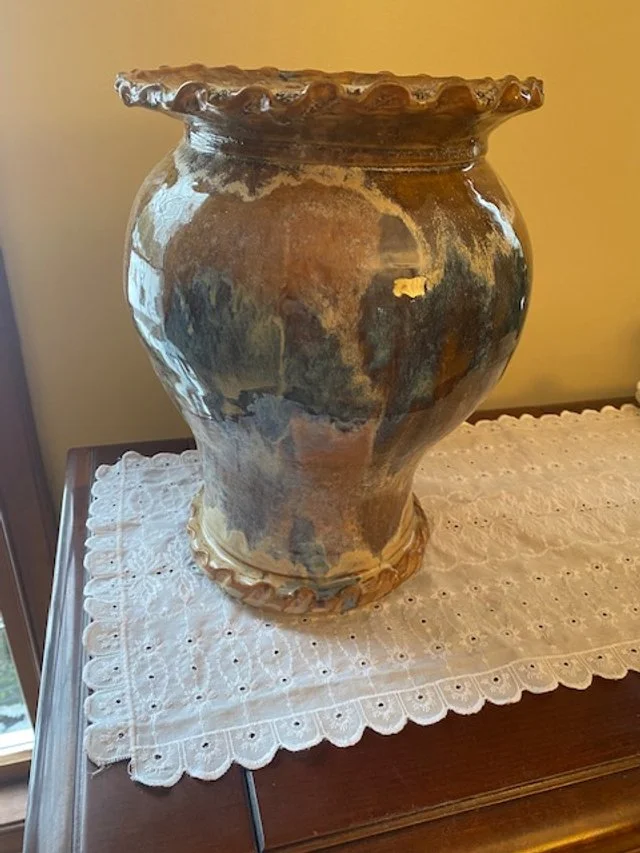 A large, decorative ceramic vase with a textured, wavy rim and a glazed, multi-colored finish, sitting on a white lace doily on a wooden surface.