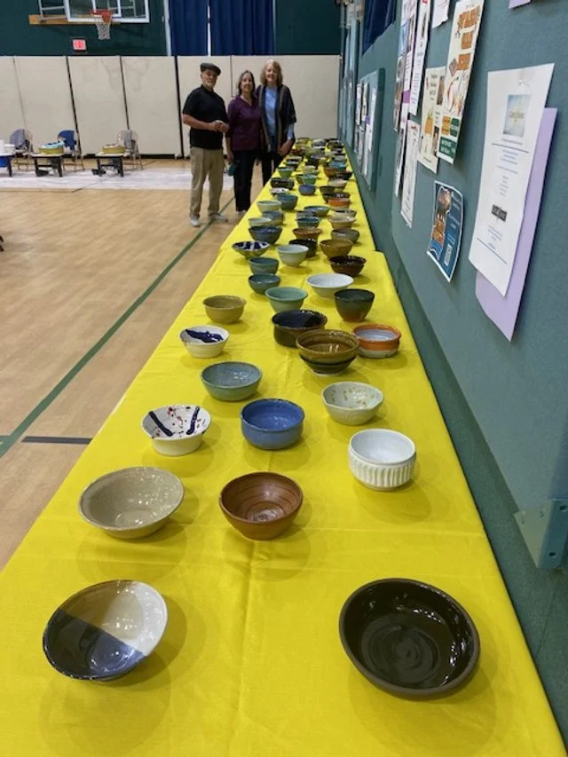 A long yellow table covered with various colorful ceramic bowls in a gymnasium or community center, with three people standing behind the table and informational posters on the wall.