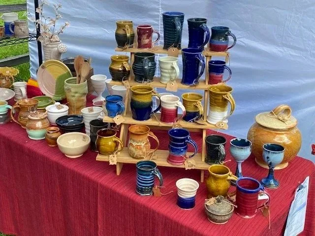 Colorful ceramic mugs, cups, bowls, and vases displayed on a table with a red tablecloth at a market stall.