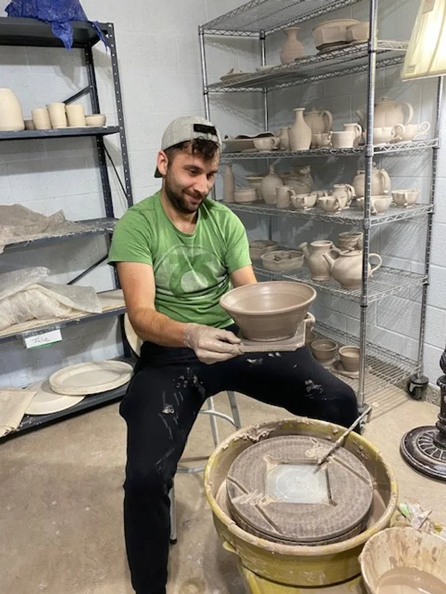 A man sitting on a stool in a pottery studio holding a finished ceramic bowl, surrounded by shelves filled with unglazed pottery.