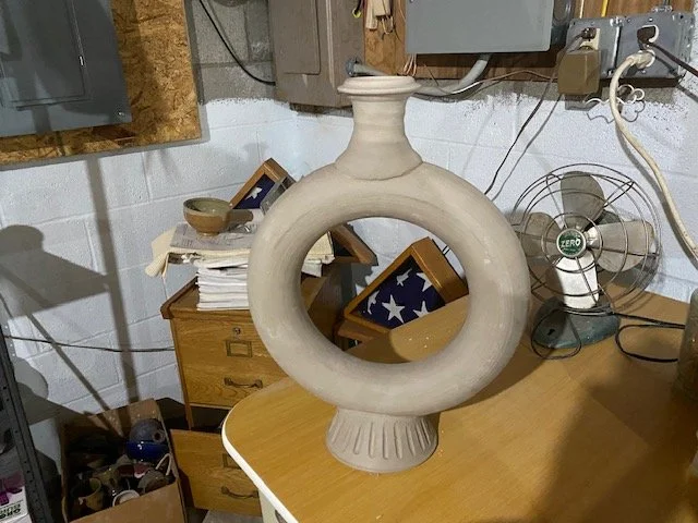 A large, beige ceramic or stone vase with a circular opening in the middle, placed on a wooden desk in a cluttered room. The room has a fan, electrical panels, and various papers and objects.