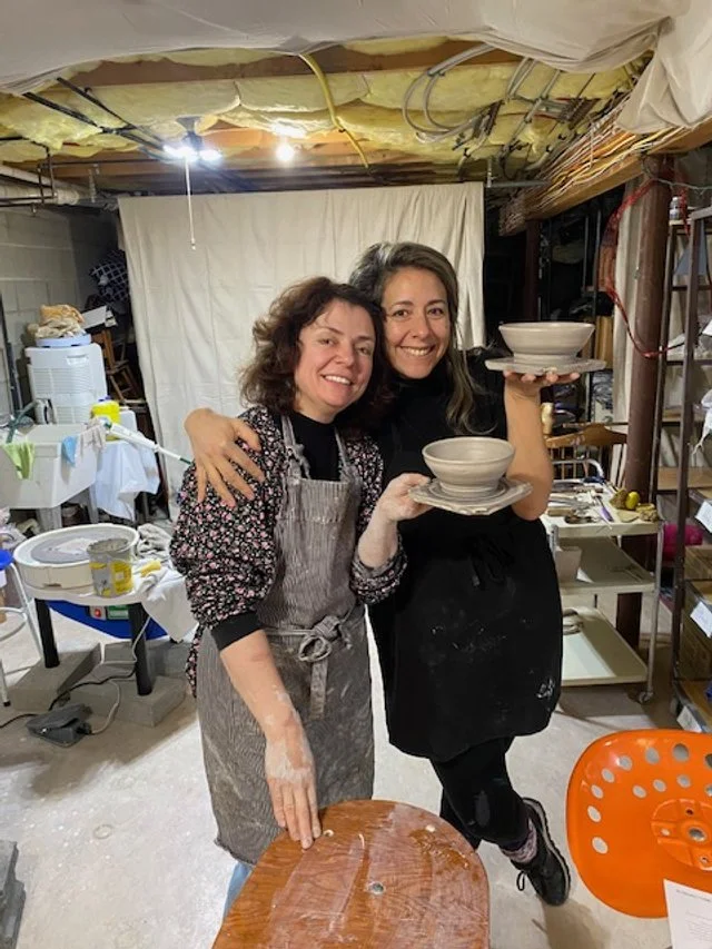 Two women smiling and holding pottery bowls in an art studio or pottery workshop.