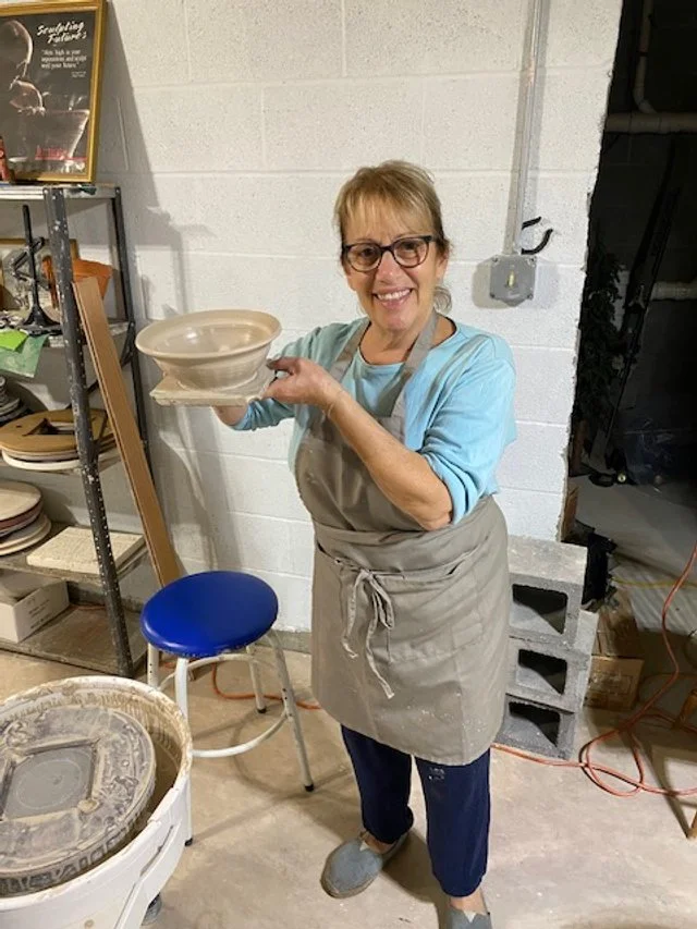 A woman with glasses and a gray apron smiling in a pottery studio, holding a finished ceramic bowl.