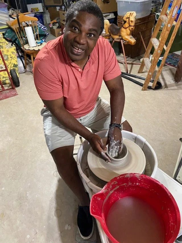 A man shaping clay on a pottery wheel in a garage or studio space, smiling at the camera.