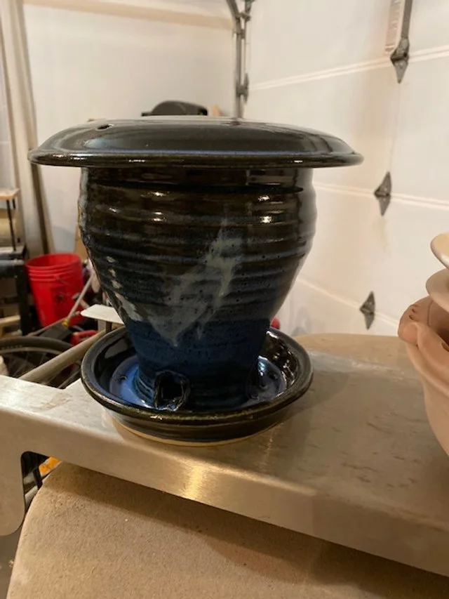 A ceramic fountain with water flowing down from the top into a basin. The fountain is black with a glossy finish, placed on a wooden surface in a garage or workshop area.