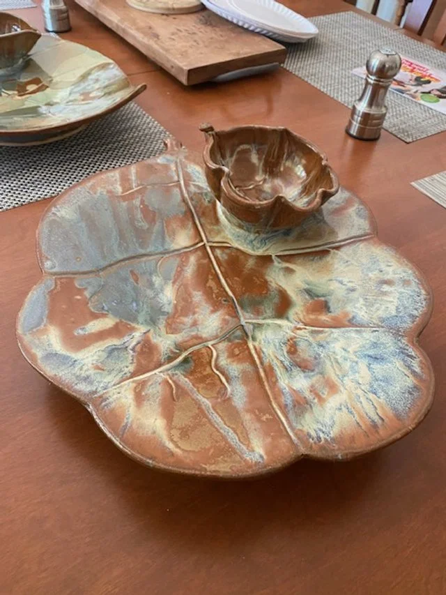 Decorative metal serving tray in the shape of a large leaf with a small bowl resembling a flower on top, placed on a wooden dining table with other dishes and utensils.