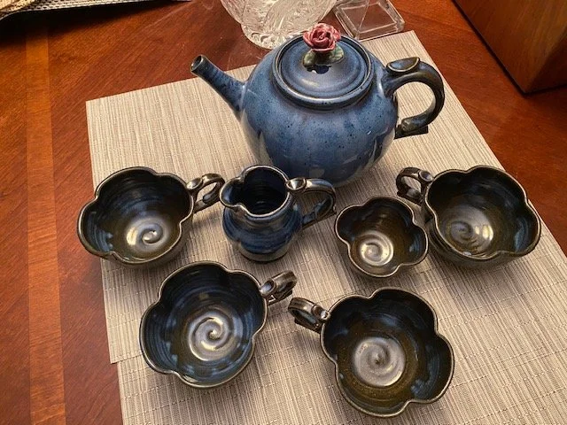 A blue teapot with a pink rose on its lid, surrounded by seven dark-colored teacups with swirl patterns on the inside, arranged on a table with a beige placemat.
