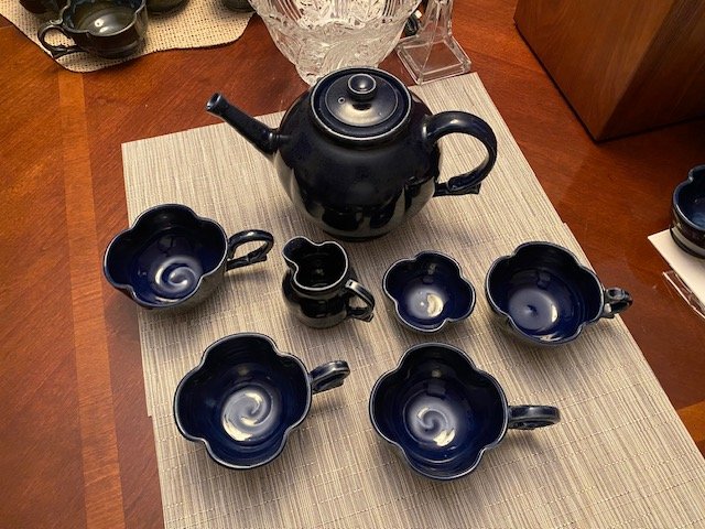 Black ceramic teapot with seven matching cups on a beige table runner